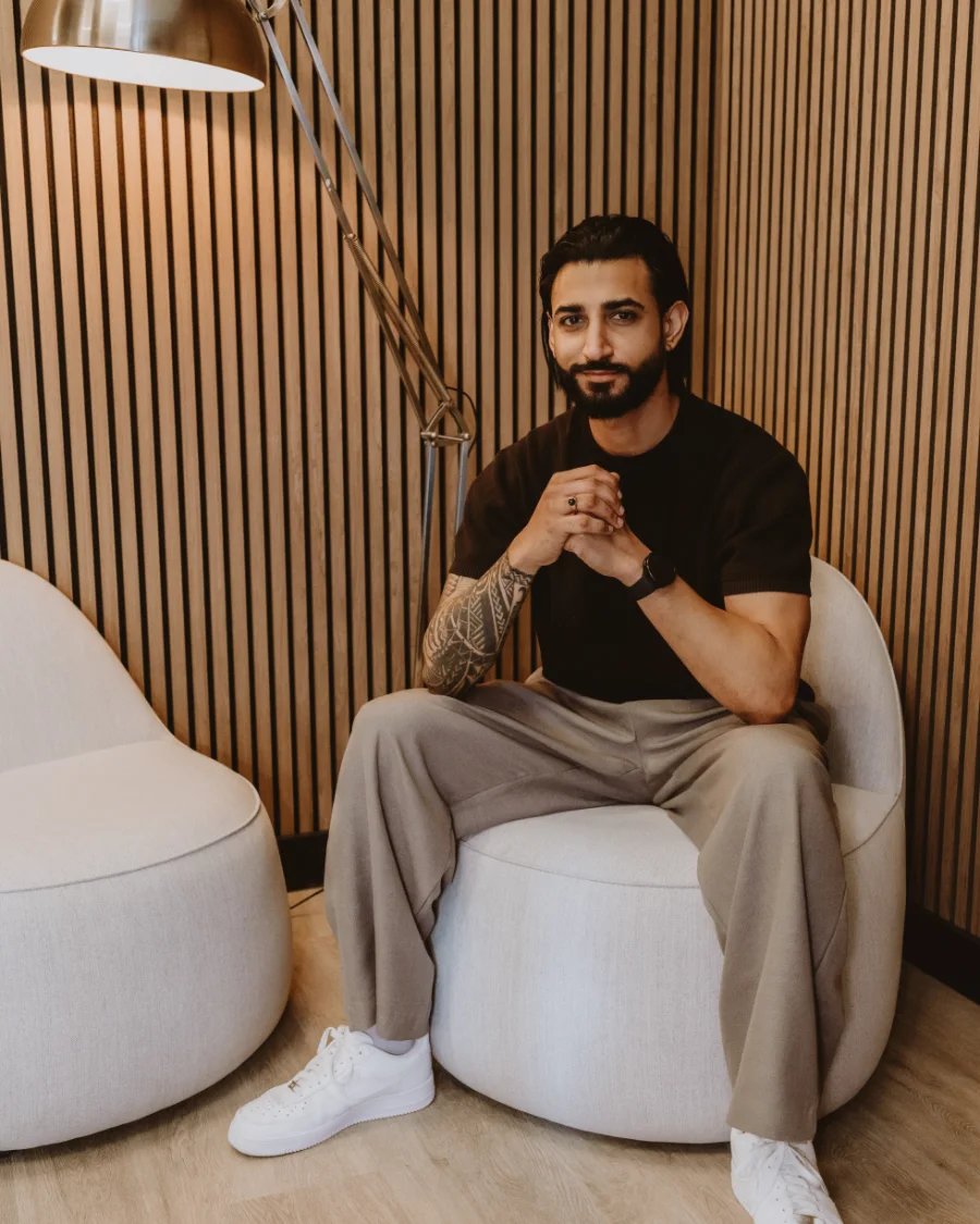 Dr Tevin Naidu in a black T-shirt, seated in a white armchair against a slatted wooden wall, smiling