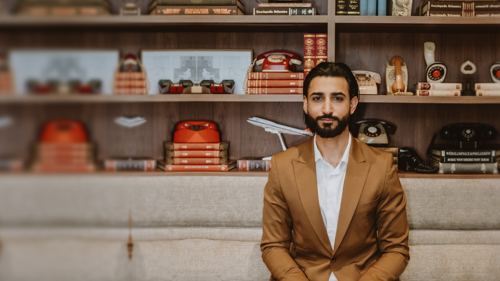 Dr Tevin Naidu in a camel suit and white shirt, seated on a linen sofa in front of walnut shelves of vintage rotary telephones and encyclopaedias.