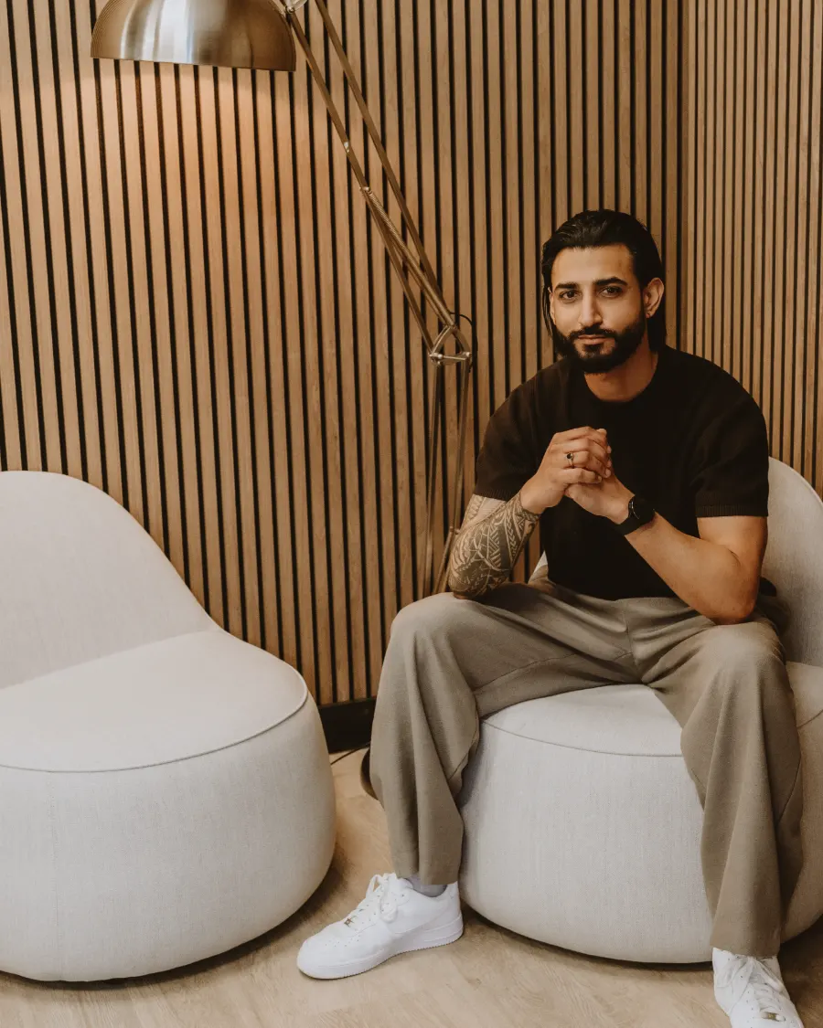 Dr Tevin Naidu in a black T-shirt, seated in a white armchair beside a brass floor lamp, hands clasped, smiling