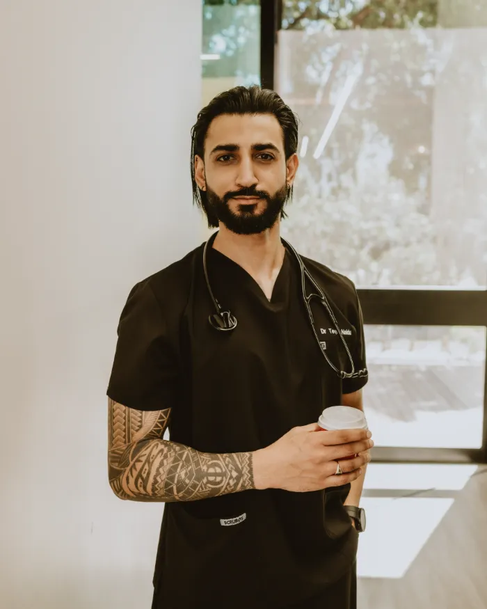 Dr Tevin Naidu in black scrubs with a stethoscope, holding a coffee beside a sunlit window