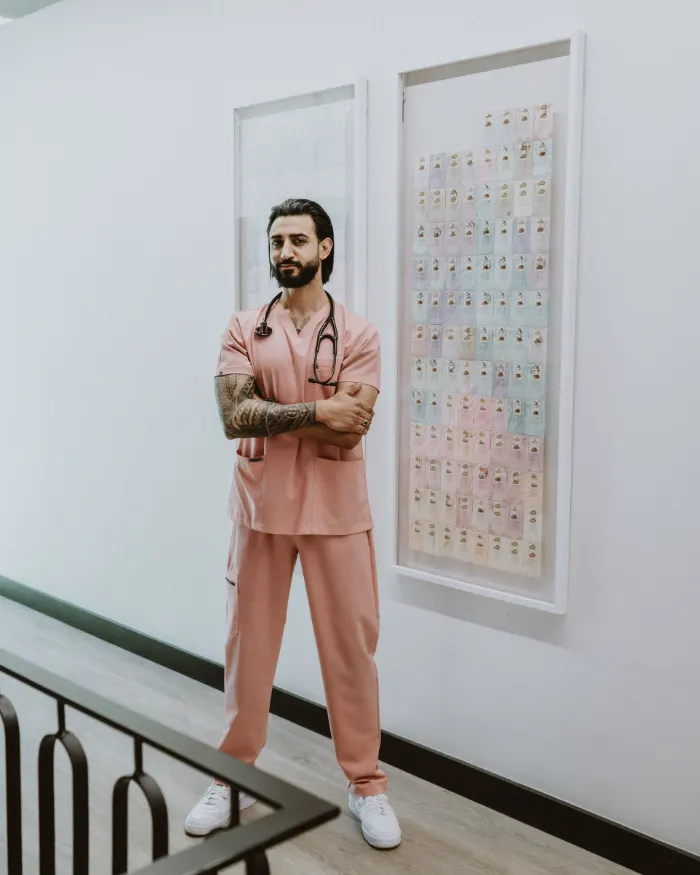 Dr Tevin Naidu in pink scrubs with a stethoscope, arms folded, standing in front of framed artwork in a bright hallway