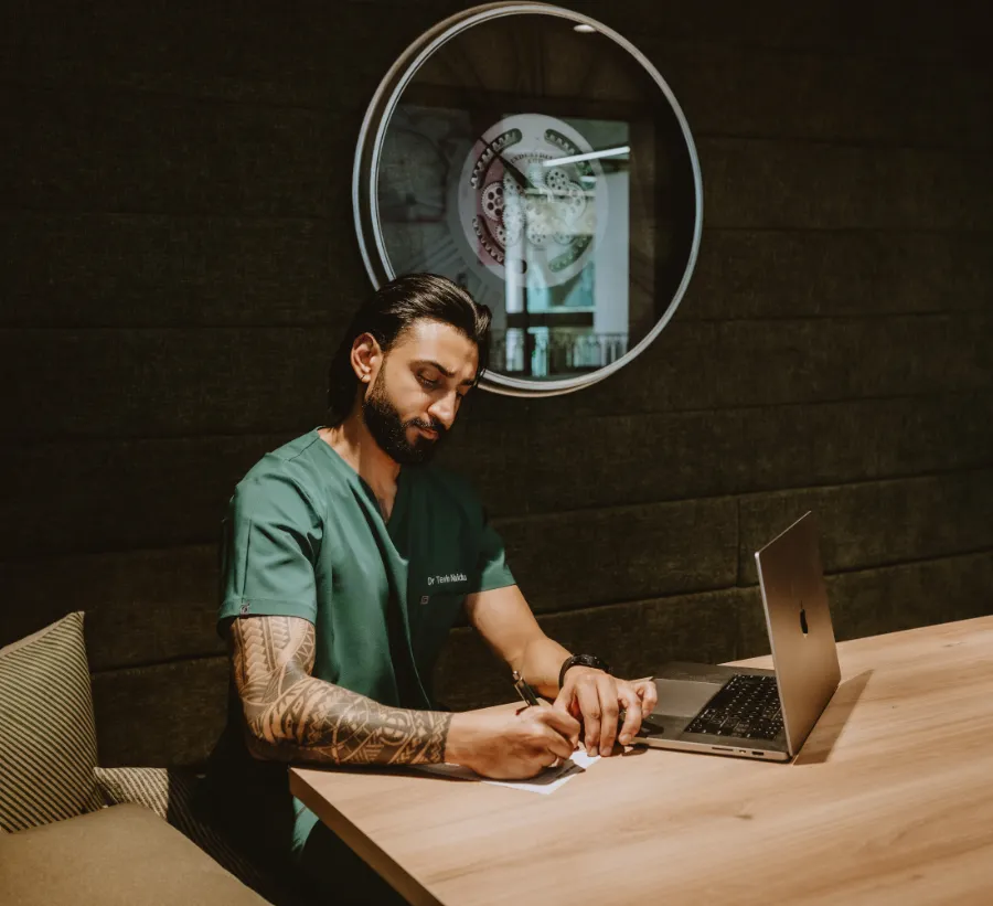 Dr Tevin Naidu in green scrubs writing notes at a consultation table beside an open laptop, a large clock on the wall behind him