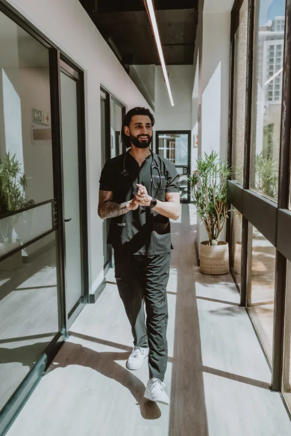 Dr Tevin Naidu in black scrubs, smiling as he walks down a sunlit corridor