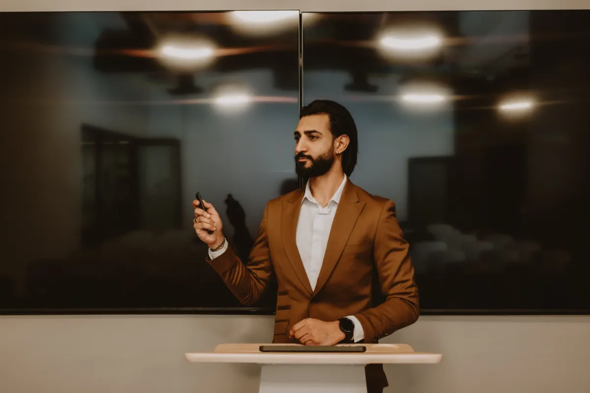 Dr Tevin Naidu presenting at a lectern in a camel suit, pointing to the screen behind him