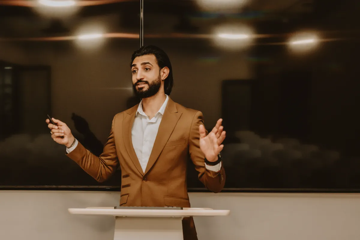 Dr Tevin Naidu speaking at a lectern in a camel suit, both hands open as he addresses the room
