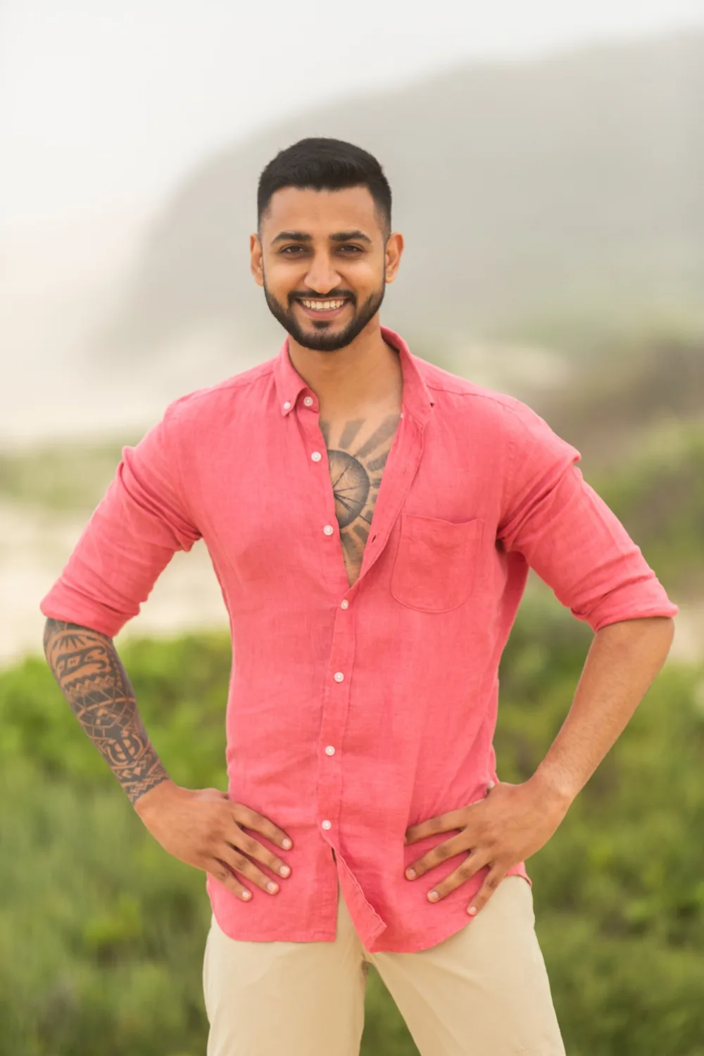 Dr Tevin Naidu smiling with his hands on his hips in a coral linen shirt, cast portrait for Survivor South Africa: Return of the Outcasts, 2022
