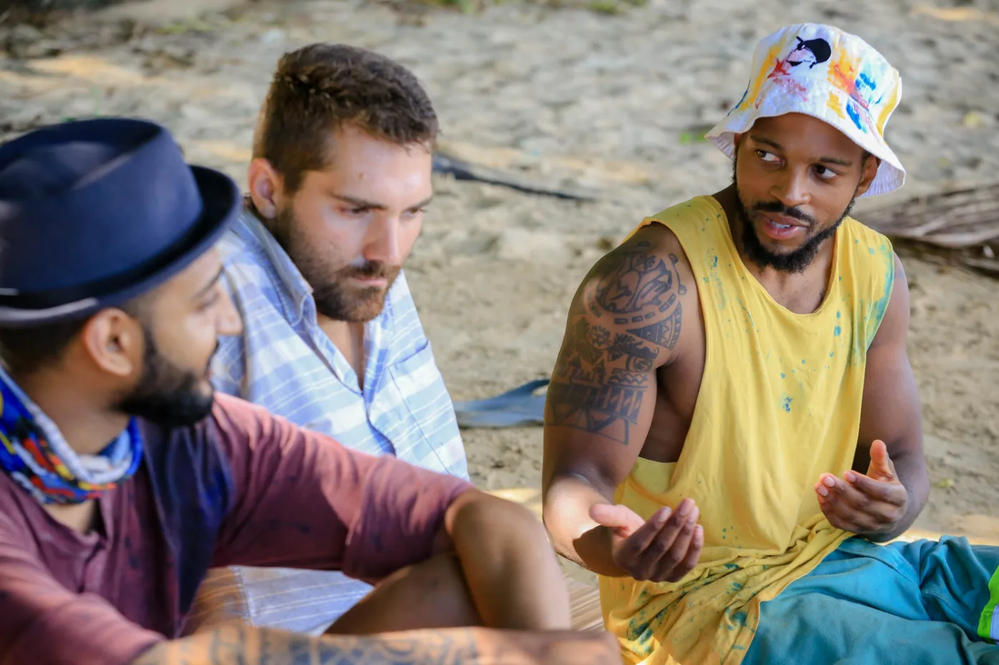 Dr Tevin Naidu in a black hat listening as a fellow Mindanao castaway talks through strategy at camp, Survivor South Africa: Philippines